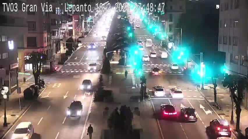 A nighttime street scene shows several cars driving through a multi-lane intersection with traffic signals, surrounded by city buildings.