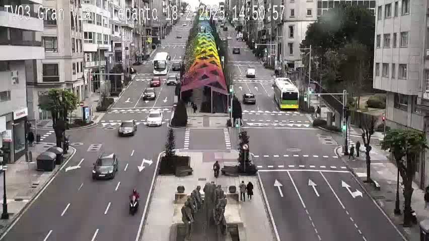 A city street scene with several cars and buses, a large colorful structure in the center, and pedestrians, on an overcast day.