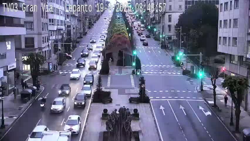 A city street scene with several cars driving in both directions under a cloudy sky, with traffic lights and pedestrians visible.