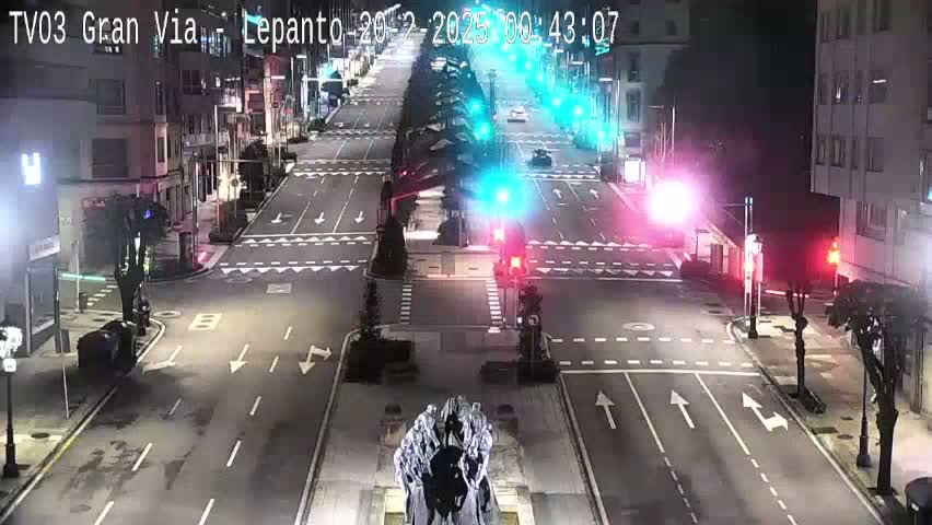 A nighttime, high-angle view of a mostly empty city intersection with traffic lights, streetlights, and a sculpture in the center.