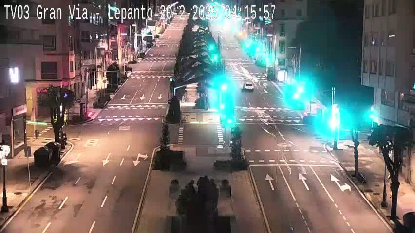 A nighttime, mostly empty city street intersection with traffic lights and a few cars visible under clear skies.