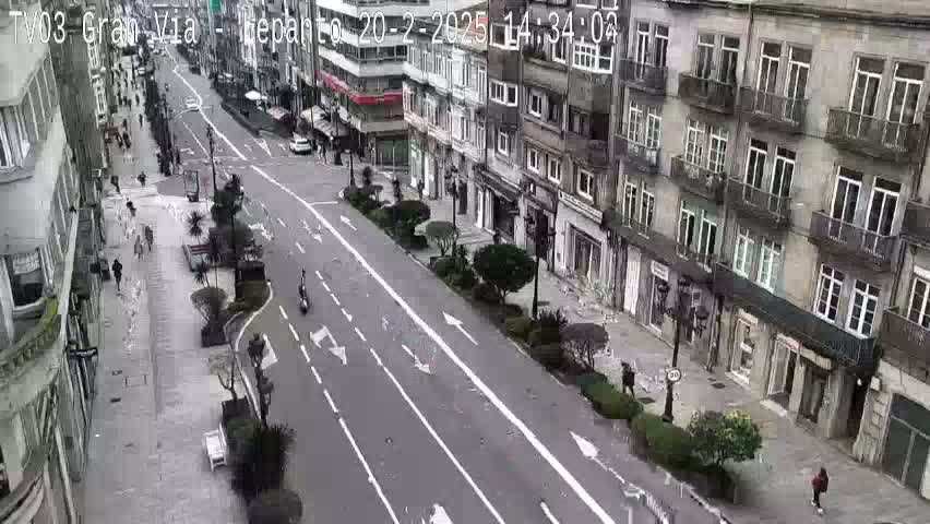 A mostly empty city street lined with tall buildings on a cloudy day, with a few pedestrians and some vehicles visible.