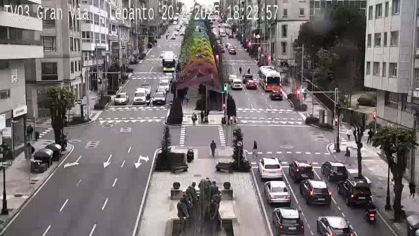 A city street scene shows overcast weather with numerous cars and buses traveling in both directions, along with pedestrians on the sidewalks and a large statue in the center of the intersection.