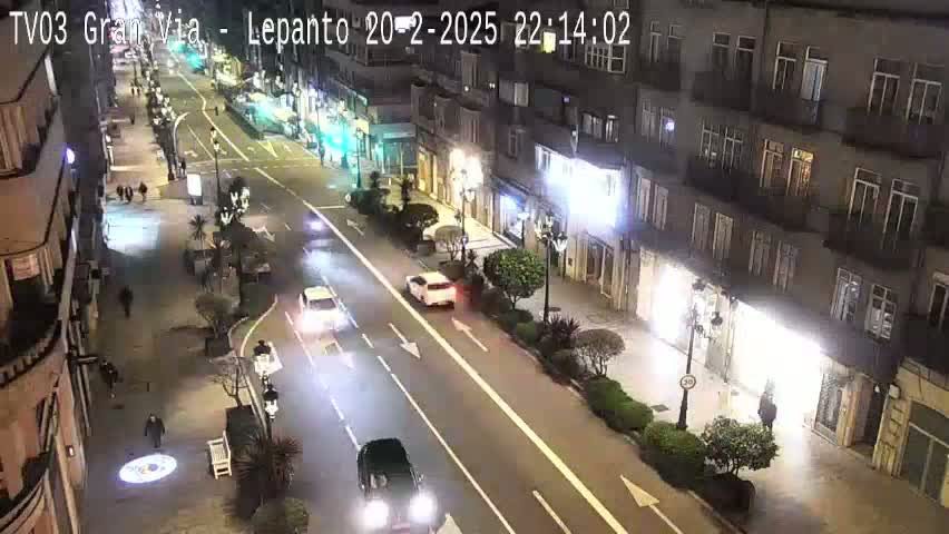 A nighttime, city street scene shows a multi-lane road with several cars, pedestrians on the sidewalk, and buildings lining both sides of the road.
