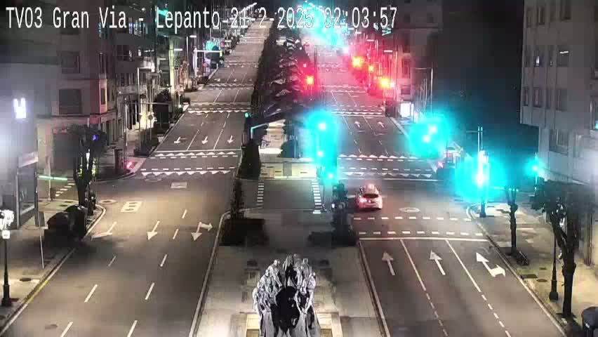 A nighttime, city street scene shows a multi-lane road with traffic lights, some vehicles, and buildings lining the sides of the road, featuring a statue in the center of the intersection.