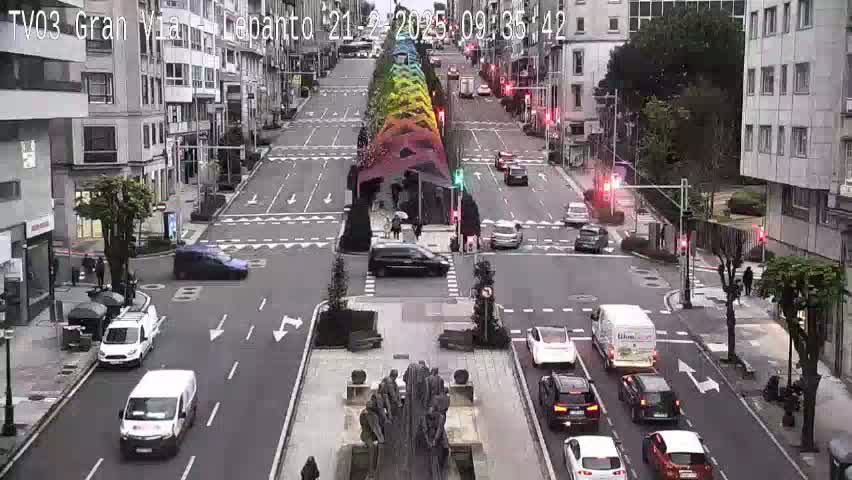 A city street scene on an overcast day shows various vehicles navigating a multi-lane roadway lined with buildings, while pedestrians walk along the sidewalks and a colorful, vertical structure stands in the center.