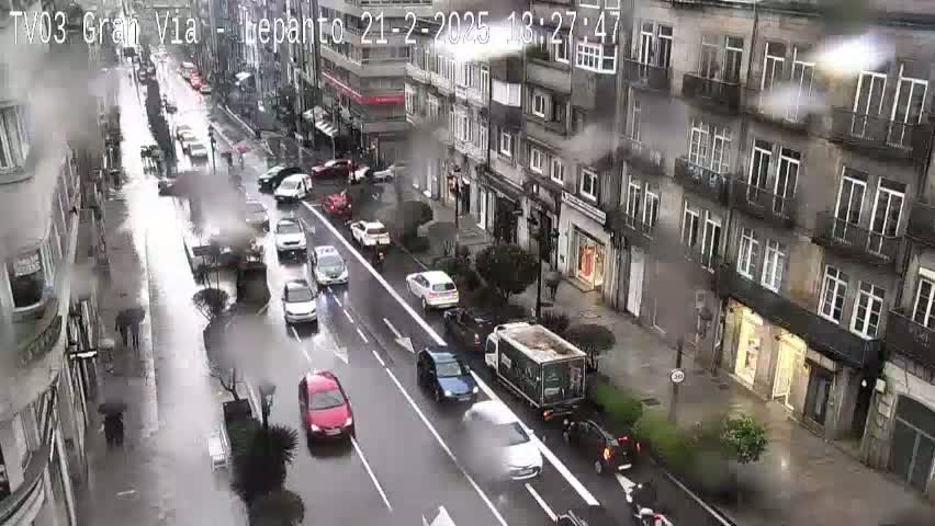 A rainy city street scene shows several cars driving on a wet road lined with multi-story buildings.