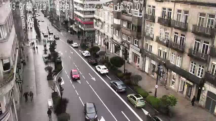 A wet city street with several cars driving down it, flanked by multi-story buildings.