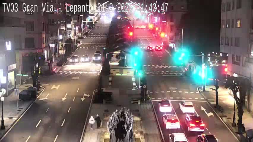 A nighttime, city street intersection with several cars and pedestrians, and traffic lights.