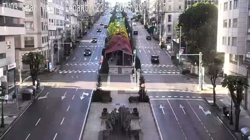 A city street scene on a sunny day shows a multi-lane road with sparse traffic, centered on a monument in a plaza, with buildings lining either side of the road.
