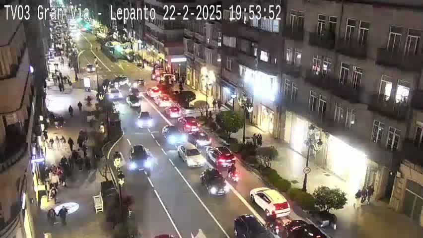 A nighttime, city street scene shows a busy road with cars moving in both directions, pedestrians walking on sidewalks next to multi-story buildings.