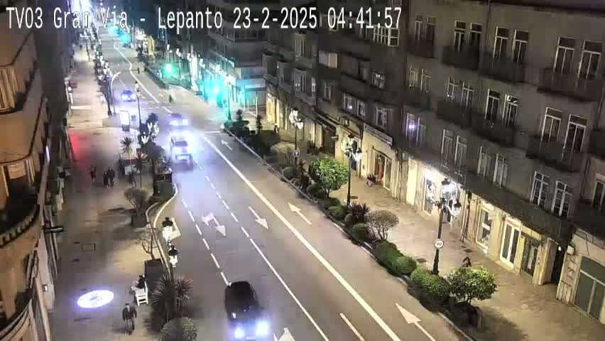 A nighttime view of a city street with several vehicles driving on a four-lane road, flanked by buildings and pedestrians.
