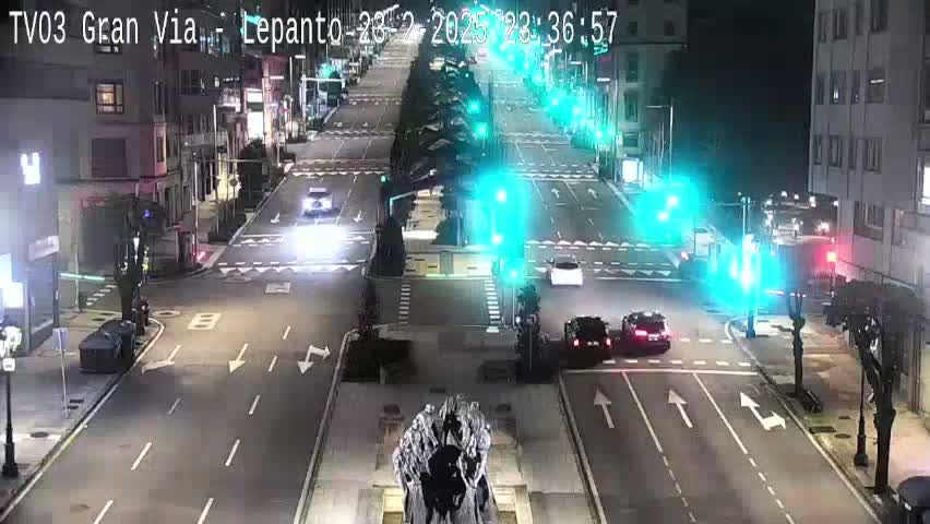 A nighttime, city street scene shows several cars at a multi-lane intersection with traffic signals, and a large statue at the center of the intersection.
