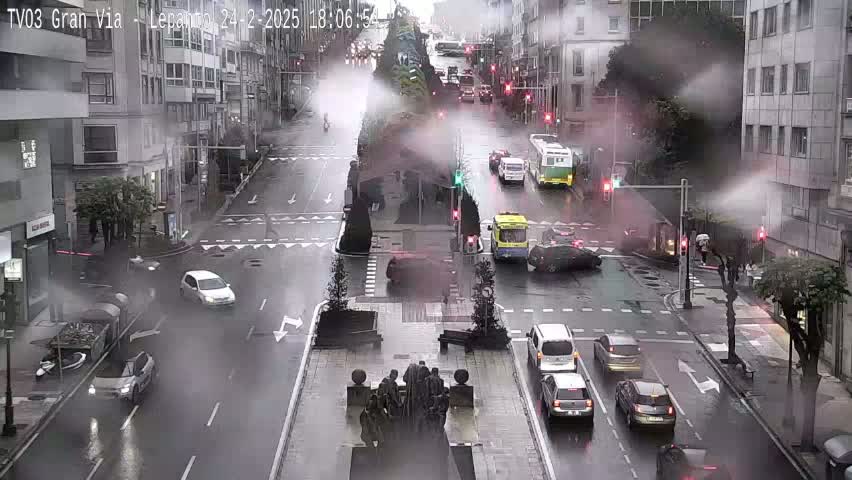 A wet city street intersection with several cars driving in the rain, and a statue in the center of the intersection.