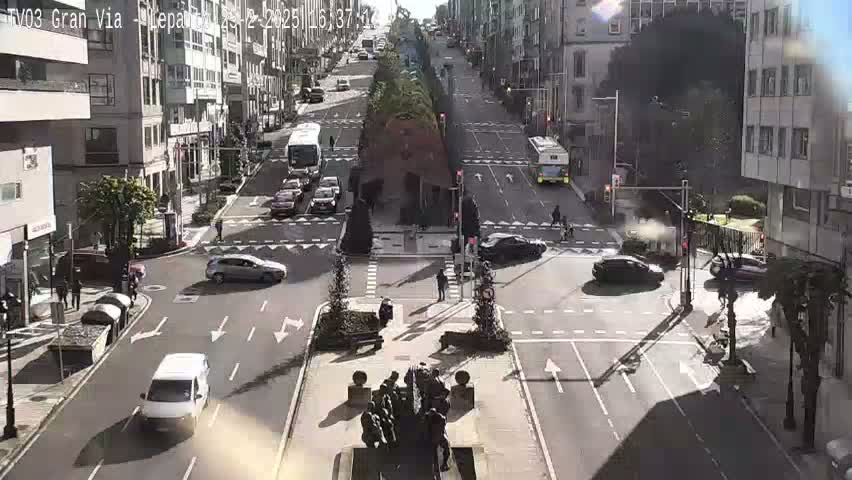 A sunny city street intersection shows several cars and buses moving in various directions, with a statue at the center of the intersection.