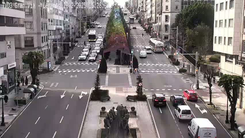 A city intersection with several cars and buses, a median with a statue, and buildings lining the street on a cloudy day.