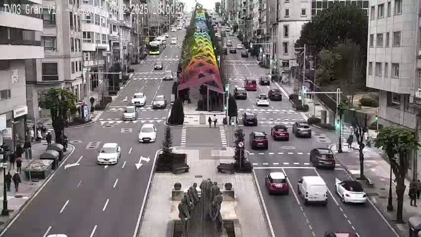 A city street scene on an overcast day shows traffic moving in both directions past a colorful, arched structure in the center, with pedestrians on sidewalks and near the crossing.