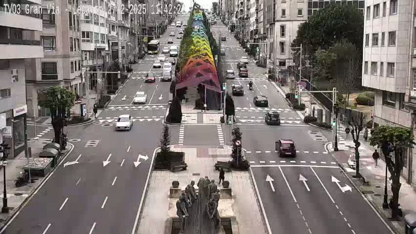 A wide city street with several cars driving in both directions under an overcast sky, features a rainbow-colored installation in the center, and a statue in the foreground.