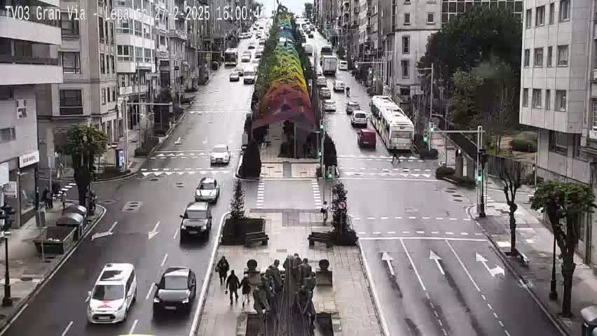 A wet city street scene shows several cars and a bus at an intersection, with a rainbow-colored structure and a statue in the center of the road.