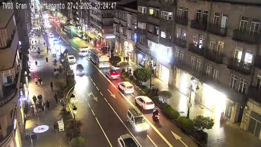 A nighttime, city street scene showing vehicles driving on a multi-lane road, flanked by pedestrians and buildings with streetlights illuminating the area.
