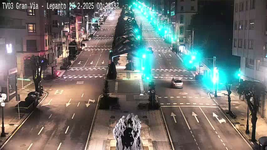 A mostly empty city street at night, with a few cars and traffic lights, features a statue in the center and buildings lining the sides under a clear sky.