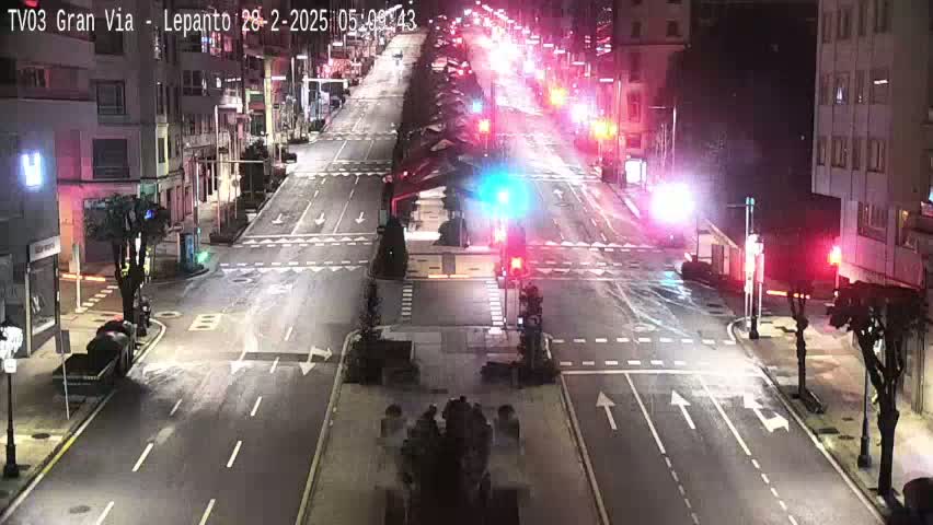 A nighttime view of a city street intersection with light traffic, wet pavement, and streetlights illuminating the scene.