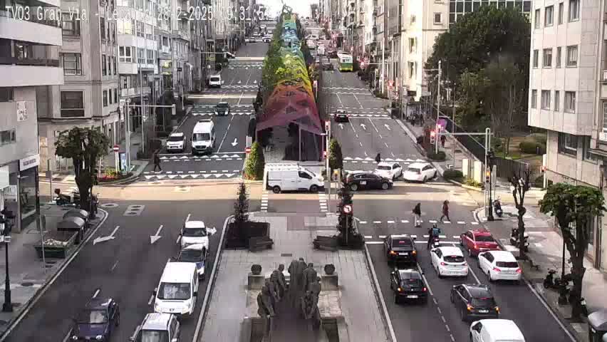 A sunny city street scene shows various cars at an intersection, with a rainbow-colored structure in the center of the road.