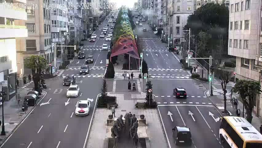 A city street scene with a few cars and pedestrians, under a mostly sunny sky, shows a monument in the center of the intersection.
