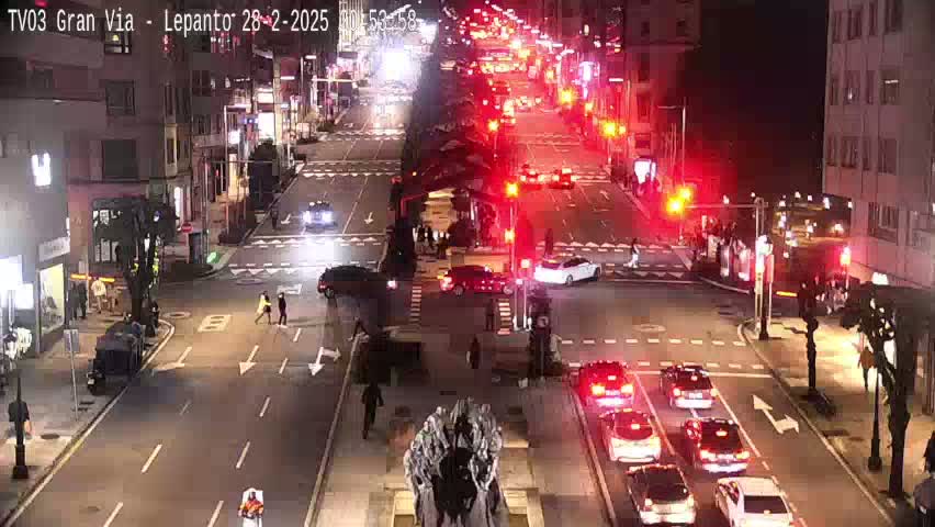A nighttime, city street scene with moderate traffic flow at an intersection, showing multiple cars and pedestrians.