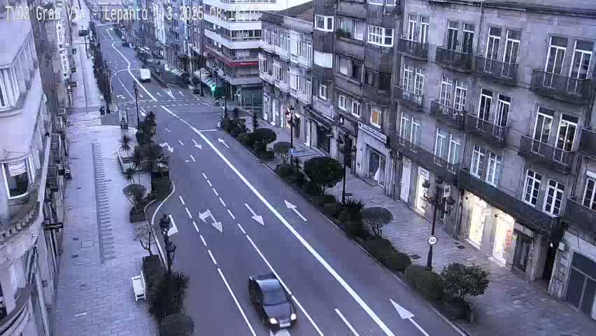 A dark-colored car drives down a city street lined with multi-story buildings under an overcast sky.