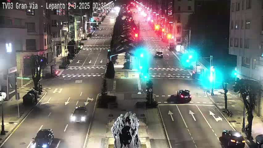 A nighttime, city street intersection with several cars, traffic lights, and a statue in the center, under clear skies.