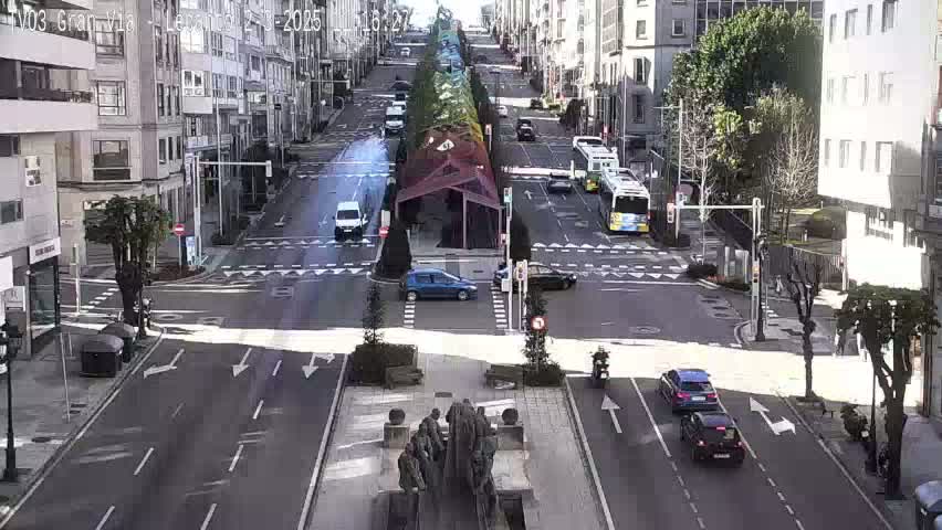 A sunny city street intersection shows several vehicles and a statue in the center, with buildings lining both sides of the street.
