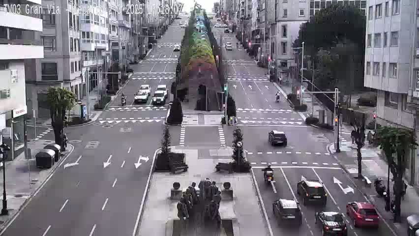 A city street intersection with several cars and pedestrians, under overcast skies, features a central monument surrounded by trees.