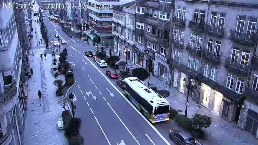 A city street scene on an overcast day shows several vehicles including a bus, and pedestrians walking along a sidewalk lined with multi-story buildings.