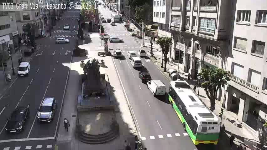 A sunny city street scene shows various cars and a bus driving on a multi-lane road that passes a statue in a plaza.