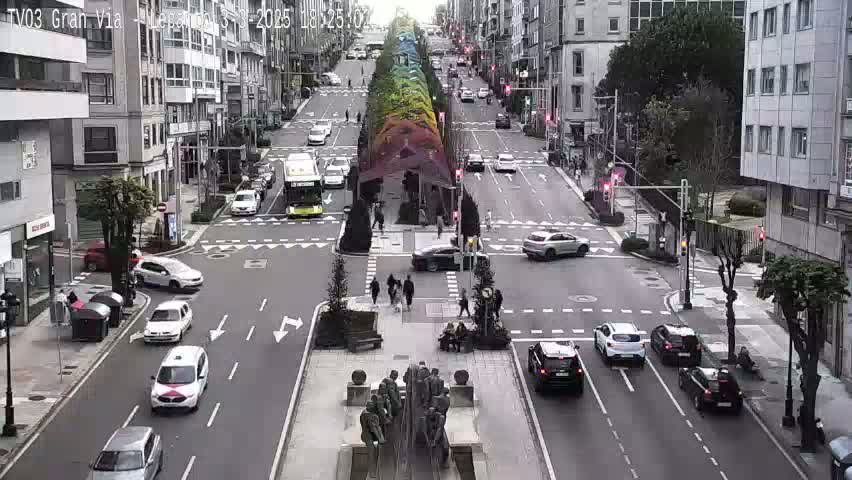 A city street scene with overcast conditions shows several cars at an intersection with pedestrians crossing and a colorful installation in the middle of the street.