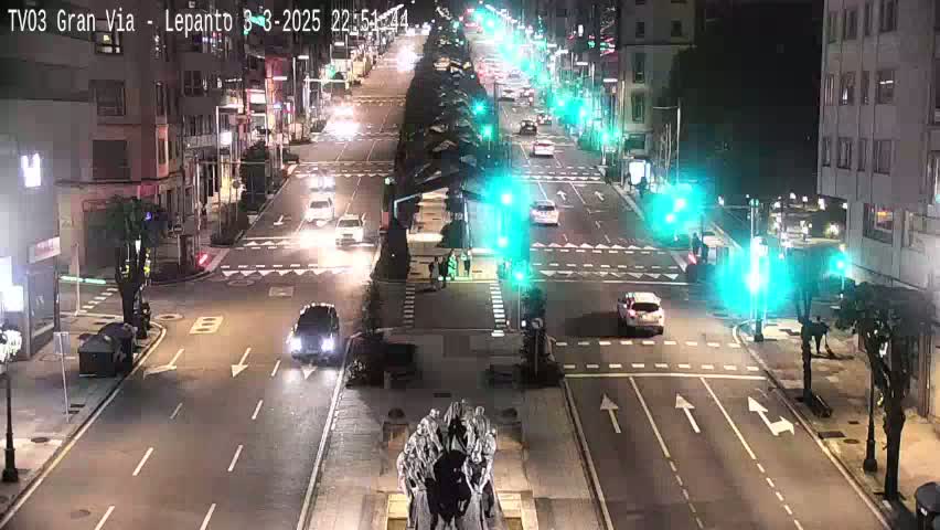 A nighttime, city street scene shows several vehicles driving along a multi-lane road, with traffic lights and streetlights illuminating the area.