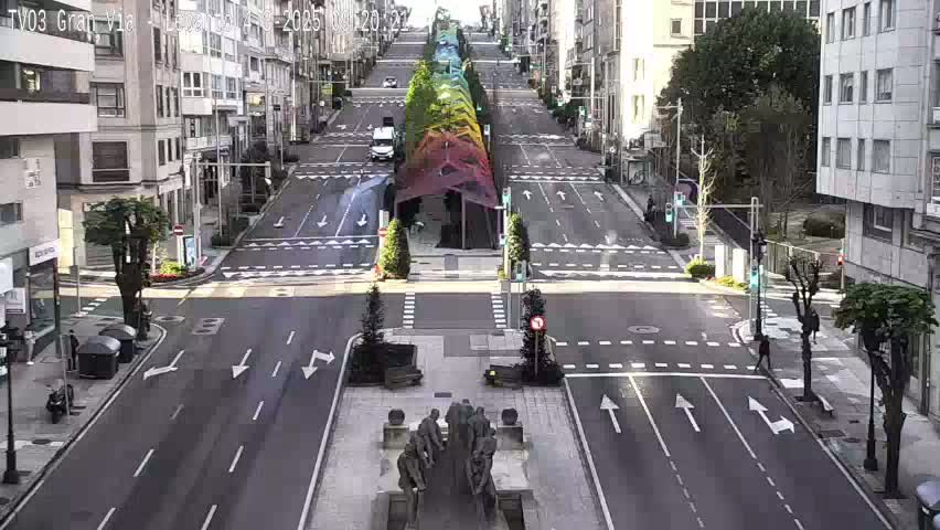 A wide, mostly empty city street intersects with a brightly colored pedestrian walkway, featuring a statue in the center, under a sunny sky.
