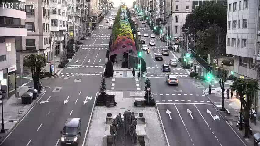 A city street scene with several cars driving on a wide road, flanked by multi-story buildings, and a central plaza with a statue; the sky is overcast.