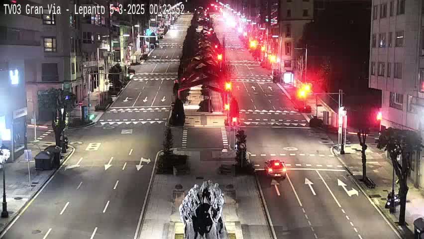 A mostly empty city street at night, illuminated by streetlights, with a few cars visible and a statue in the center of the intersection.