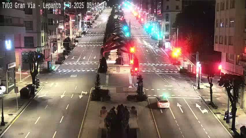 A mostly empty city street at night, with a few cars and people, is lit by streetlights and traffic signals.