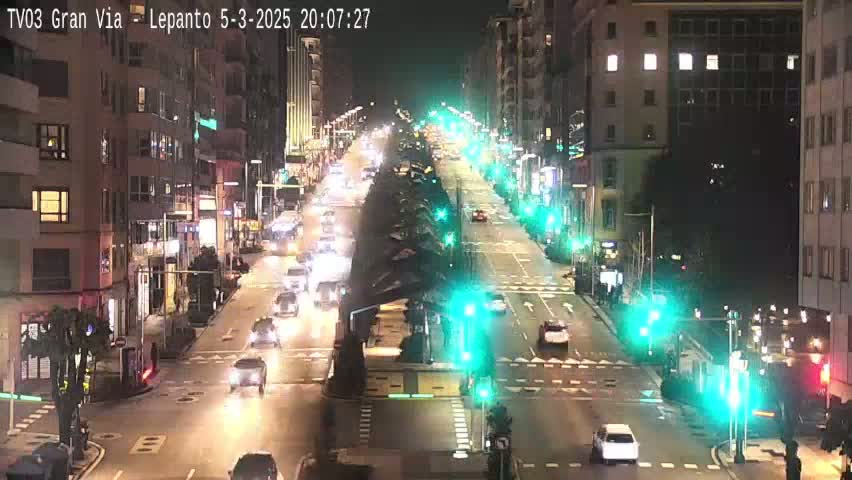 A nighttime street scene shows a multi-lane road with cars driving in both directions, lined with buildings on either side, under bright streetlights.