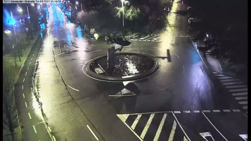 A wet, nighttime view of a roundabout with a central fountain, surrounded by roads and pedestrian crossings.