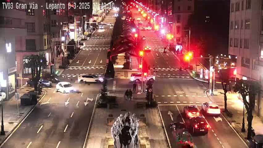 A nighttime aerial view of a city street intersection with several cars and pedestrians, under clear skies.
