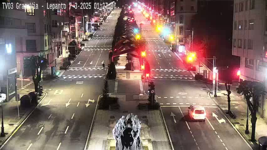 A mostly empty, wide city street at night, illuminated by streetlights, with a few vehicles and traffic signals visible.