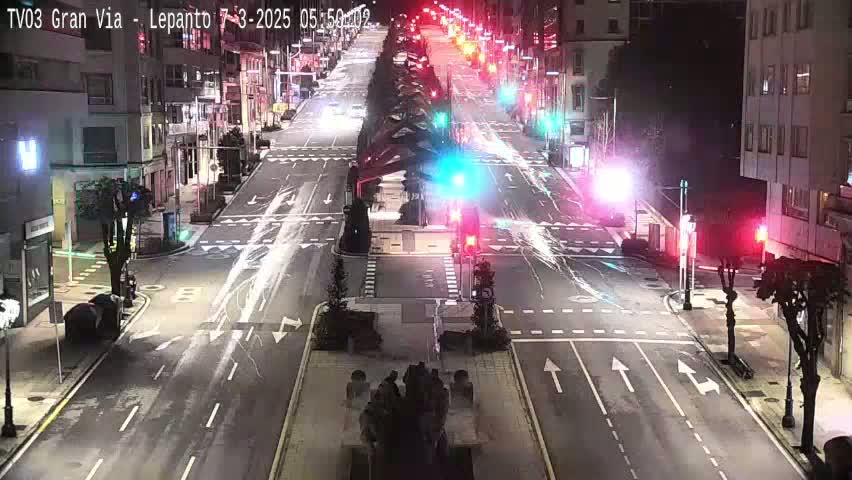 A mostly empty, multi-lane city street at night, illuminated by streetlights and traffic signals, with some cars visible in the distance.