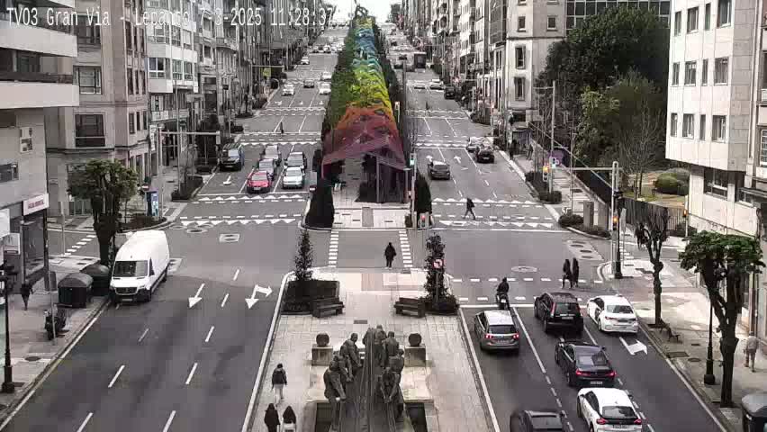 A city street scene on an overcast day shows several vehicles driving along a wide road that intersects with another road, with a large sculpture in the center of the intersection and pedestrians crossing the street.