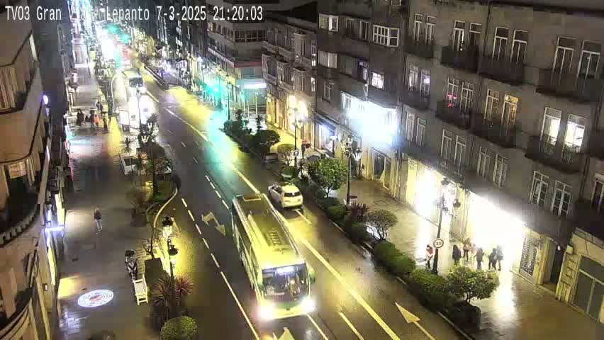 A wet city street at night, showing a bus and several cars, with pedestrians and buildings lining both sides of the street.