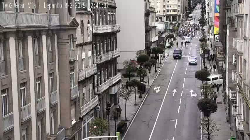 A street scene shows several people walking along a city street lined with tall buildings, with several vehicles traveling down the road, under overcast skies.