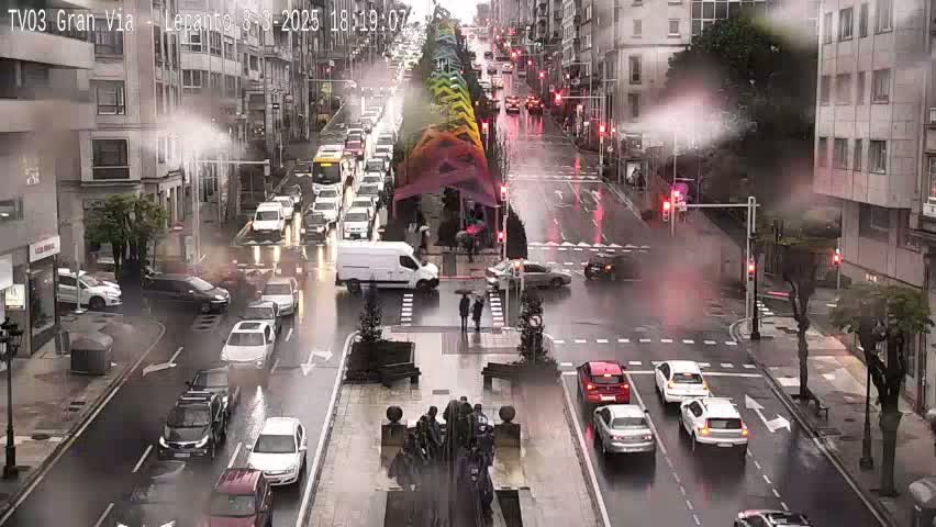 A rainy city street scene shows numerous cars driving in both directions, passing a central fountain and pedestrian crossing, with buildings lining either side of the street.